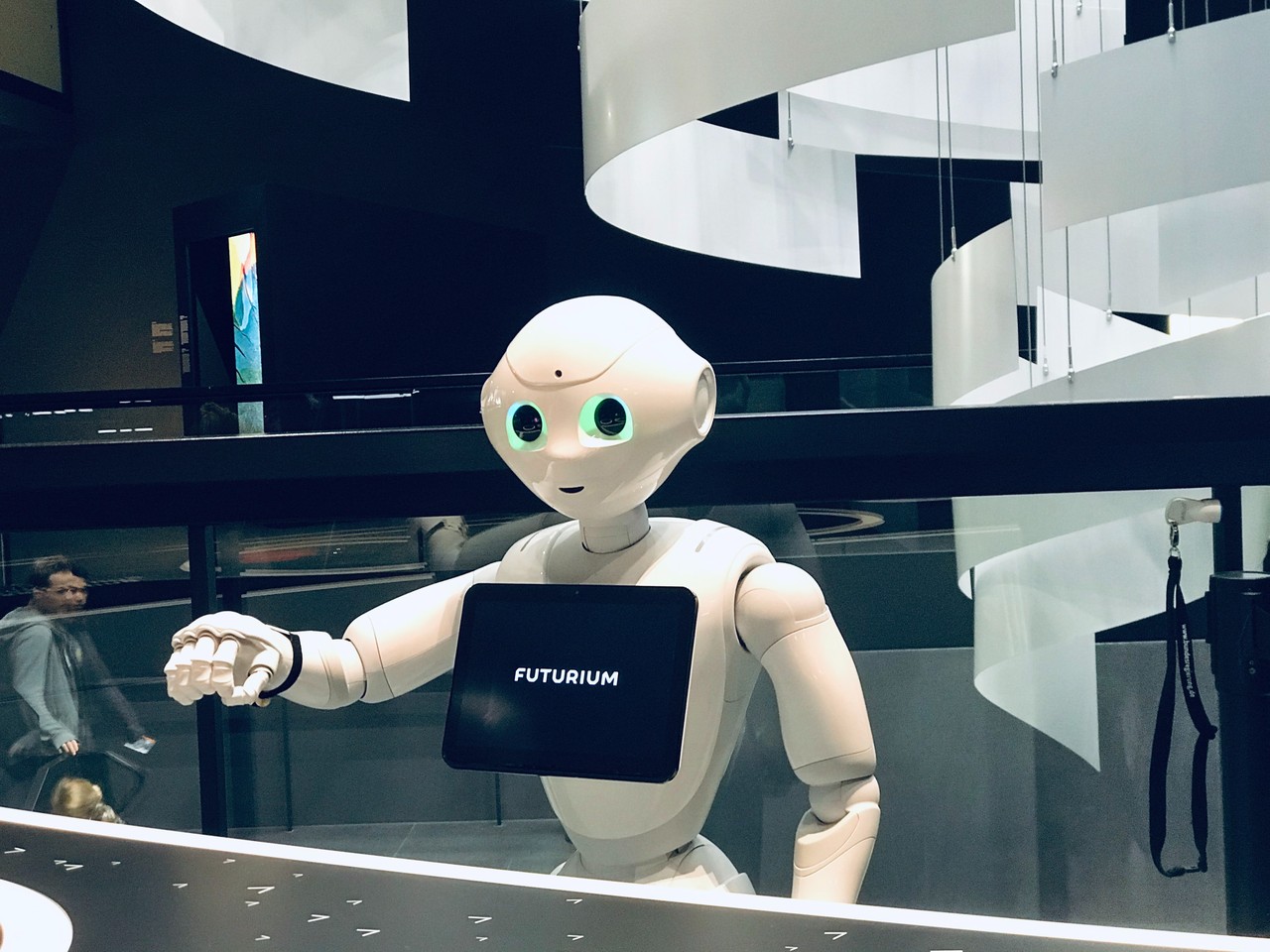 Zu sehen ist ein Roboter mit einem Tablet an seiner Vorderseite, der seinen rechten Arm ausstreckt.