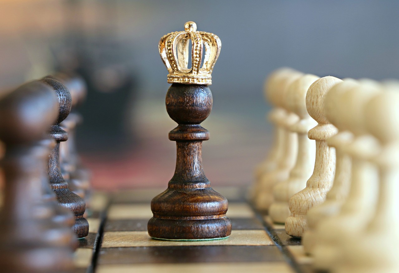 Es wird ein Schachbrett aus einer seitlichen Perspektive gezeigt. In der Mitte steht eine Schachfigur, die eine Krone trägt. Weitere Figuren stehen am Rand.