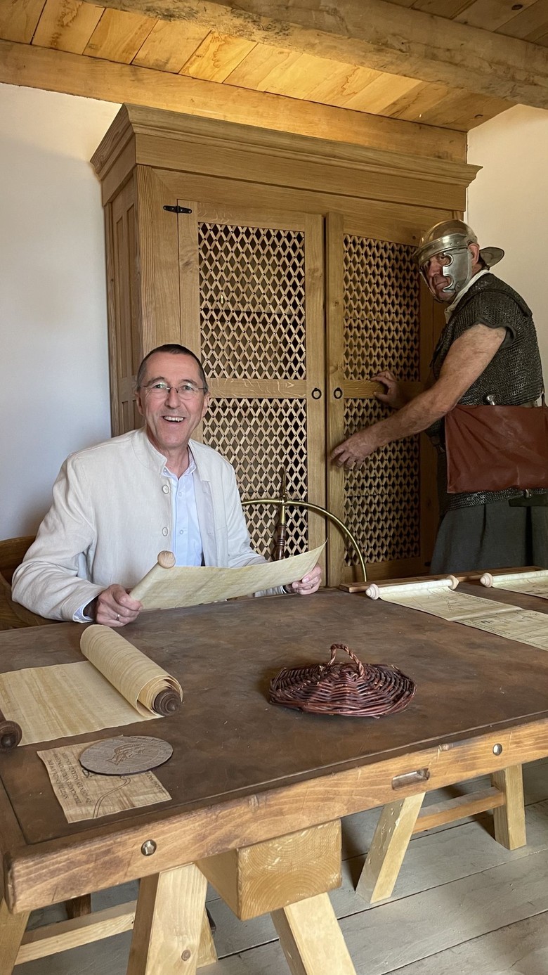Ein Mann sitzt an einem Tisch und hält eine Schriftrolle in der Hand; im Hintergrund steht ein Mann. Beide tragen antike Kleidung.