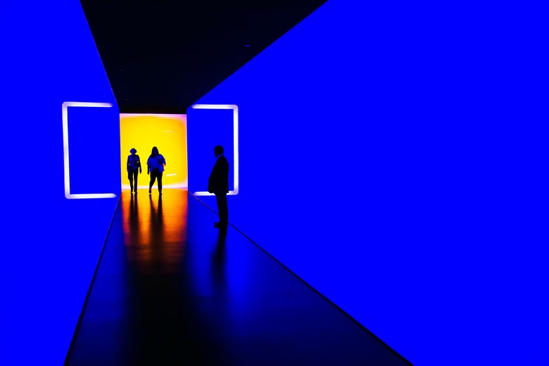 Schemen von drei Personen stehen in einem Gang, der mit gelben und blauen Lichteffekten spielt.