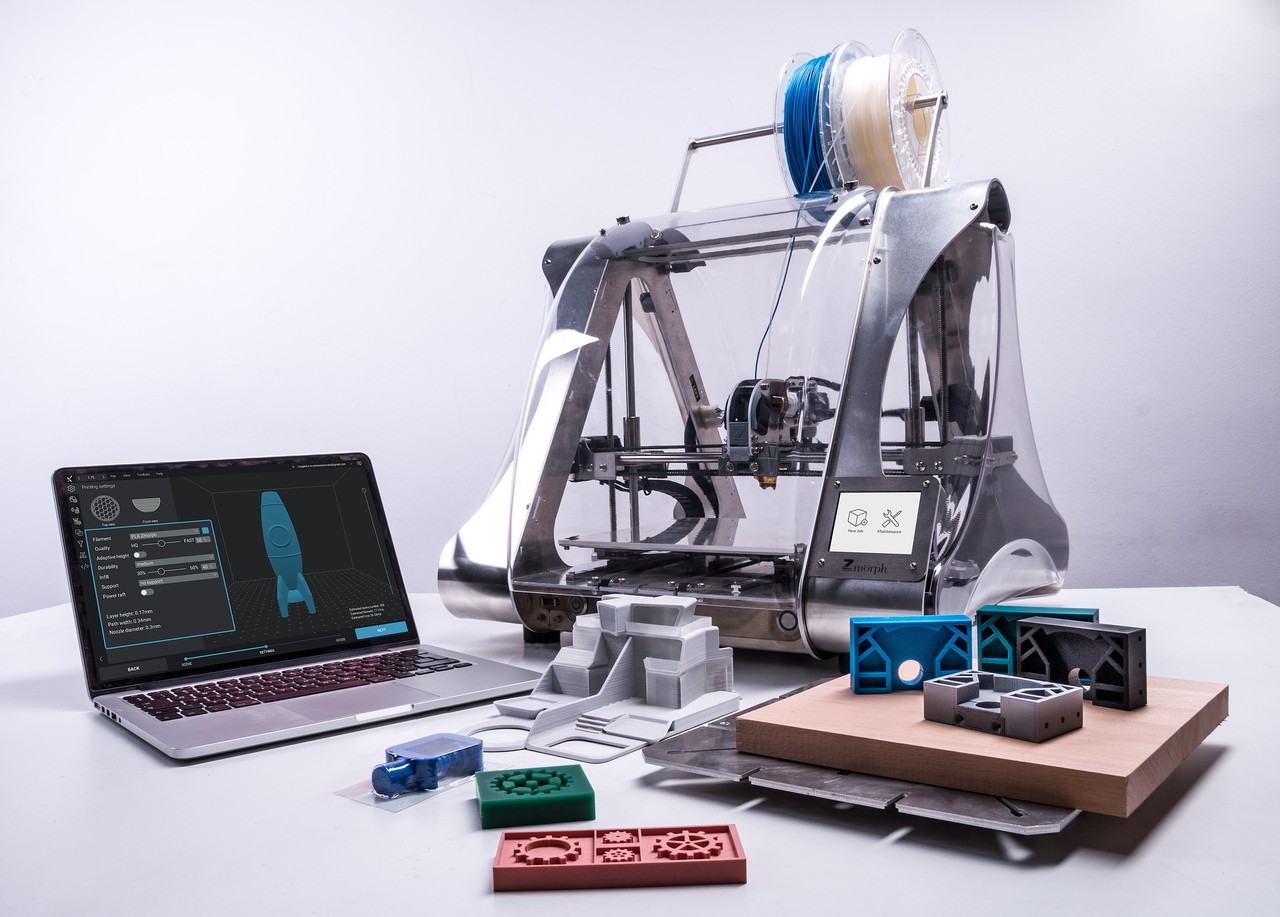 Neben einem geöffnetem Laptop steht ein 3D-Drucker, während im Vordergrund fertig gedruckte Modelle liegen.