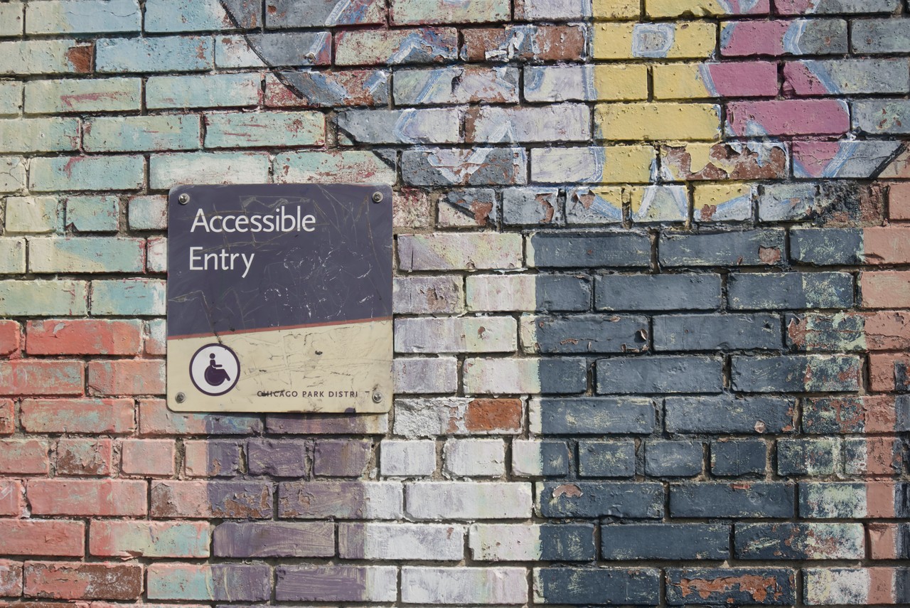 An einer bunt bemalten Mauer hängt ein schon etwas kaputtes Schild mit der Aufschrift "Accessible Entry" und einem Rollstuhlfahrer-Zeichen.